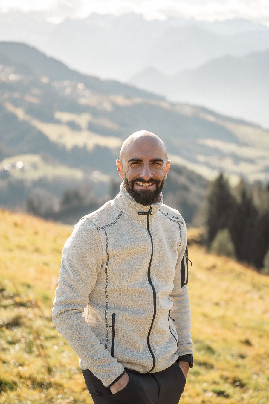 Ein Mann im Profil mit Bart und Glatze, trägt eine grüne Kapuzenjacke, sitzt auf einer Holzbank und blickt nachdenklich zur Seite.