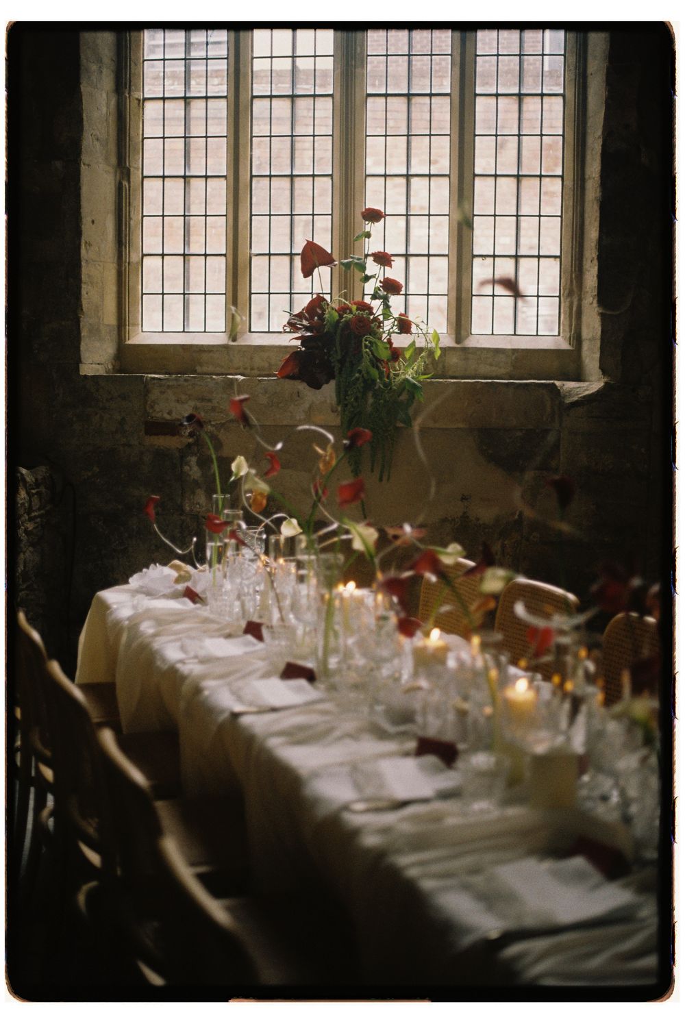 Elegant dining table set with white linens and floral centerpieces beside large windows in a rustic interior space.