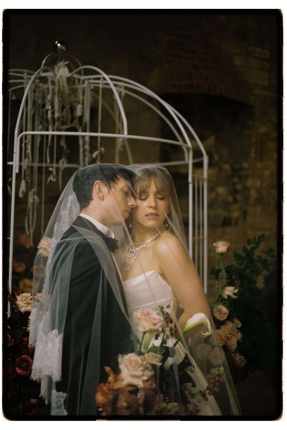 A romantic wedding couple shares an intimate moment under a decorative white arbor surrounded by soft floral arrangements.