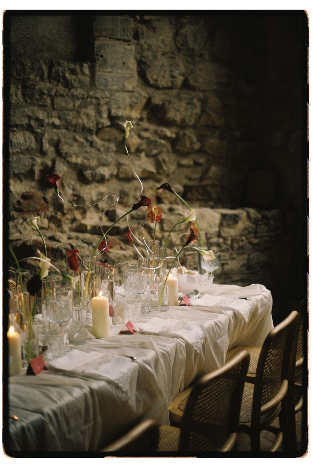Elegant dining table set with white linens, candles and glassware against a rustic stone wall in dim lighting.