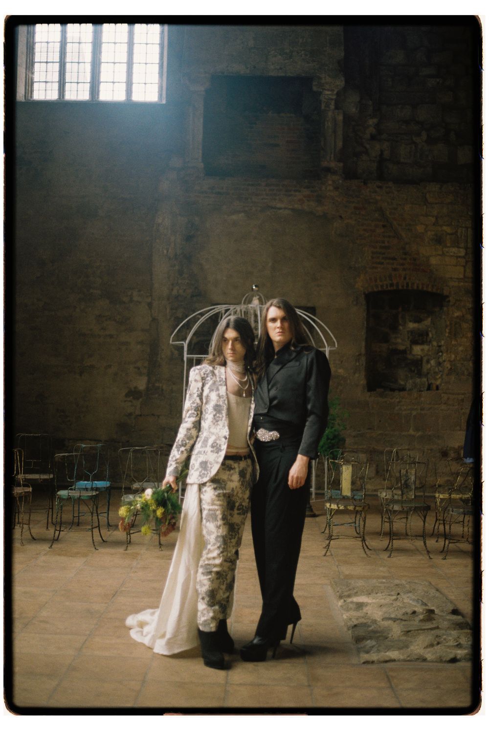 A couple in wedding attire poses in a rustic brick room with high windows and scattered chairs.