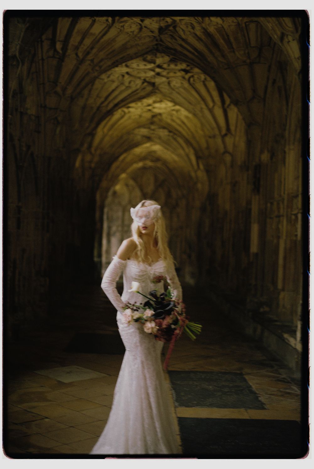 A figure in a vintage white wedding dress stands in a dimly lit stone corridor holding a bouquet of flowers.