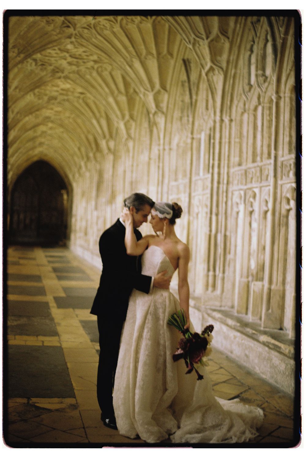 A romantic wedding couple embraces in a historic stone corridor with Gothic arched ceilings and dramatic lighting.