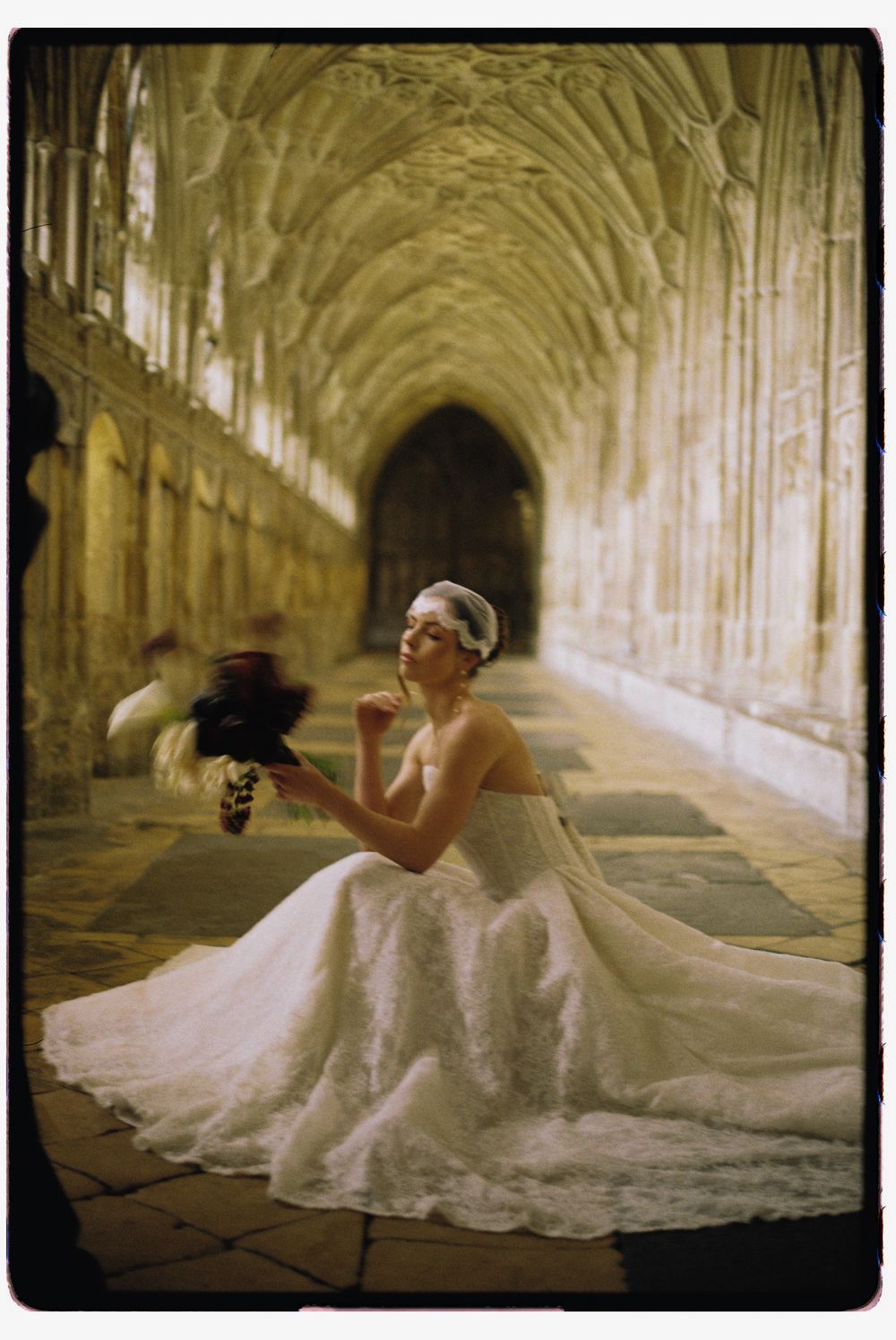 A bride in a flowing white gown sits gracefully in a gothic cathedral hallway with vaulted ceilings and stone architecture.