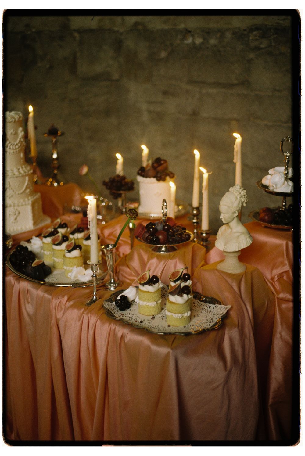 Elegant dessert table with lit candles and treats arranged on a peach-colored tablecloth in warm, moody lighting.