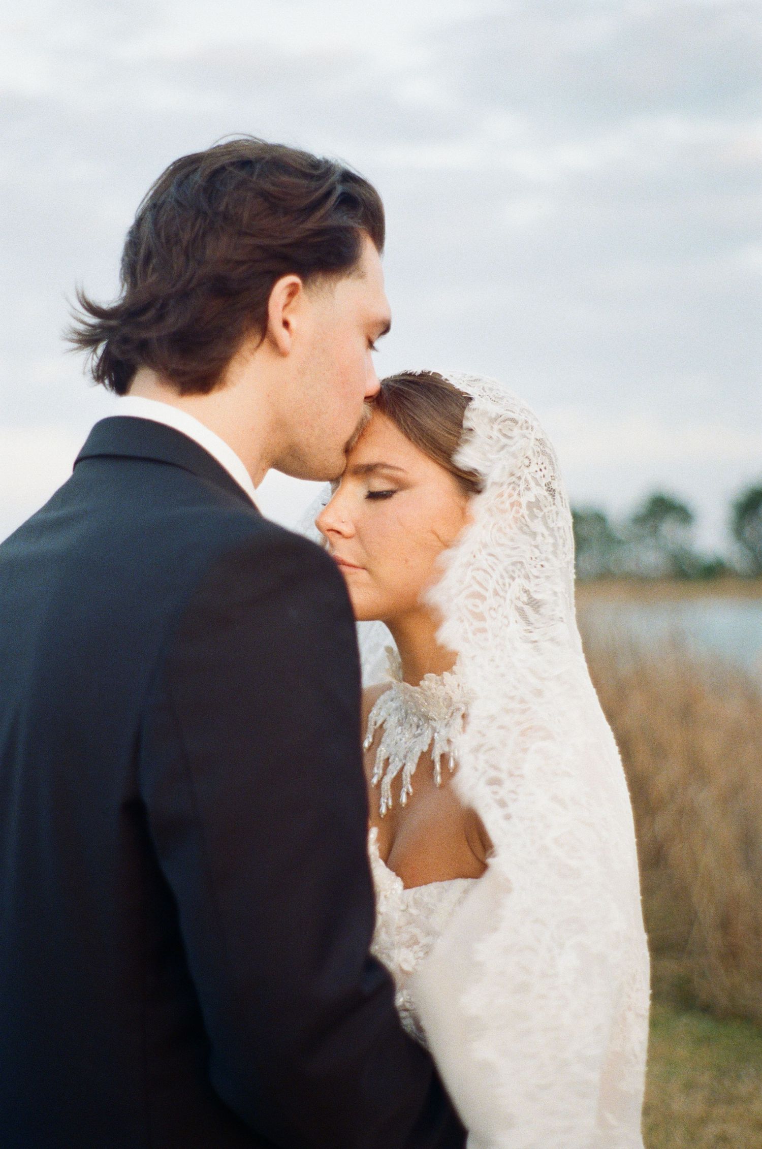 Blurry romantic photo of bride and groom walking toward golden sunset by a lake, wedding dress flowing in the breeze.
