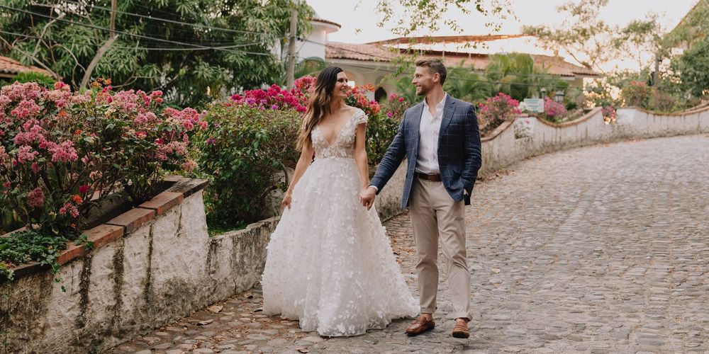 Bride in floral lace gown and groom in blue blazer walking hand in hand on cobblestone street with pink flowers.