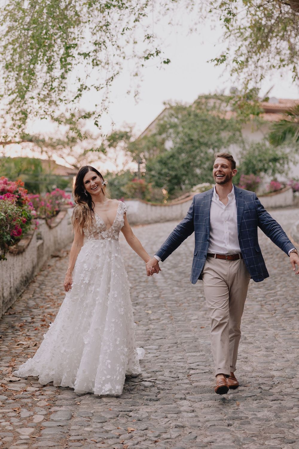 Happy bride and groom holding hands and walking on charming cobblestone street surrounded by colorful blooming flowers.