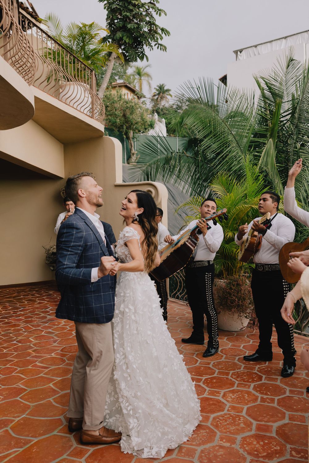 Bride and groom dancing joyfully on terracotta patio while mariachi band performs at tropical wedding venue.