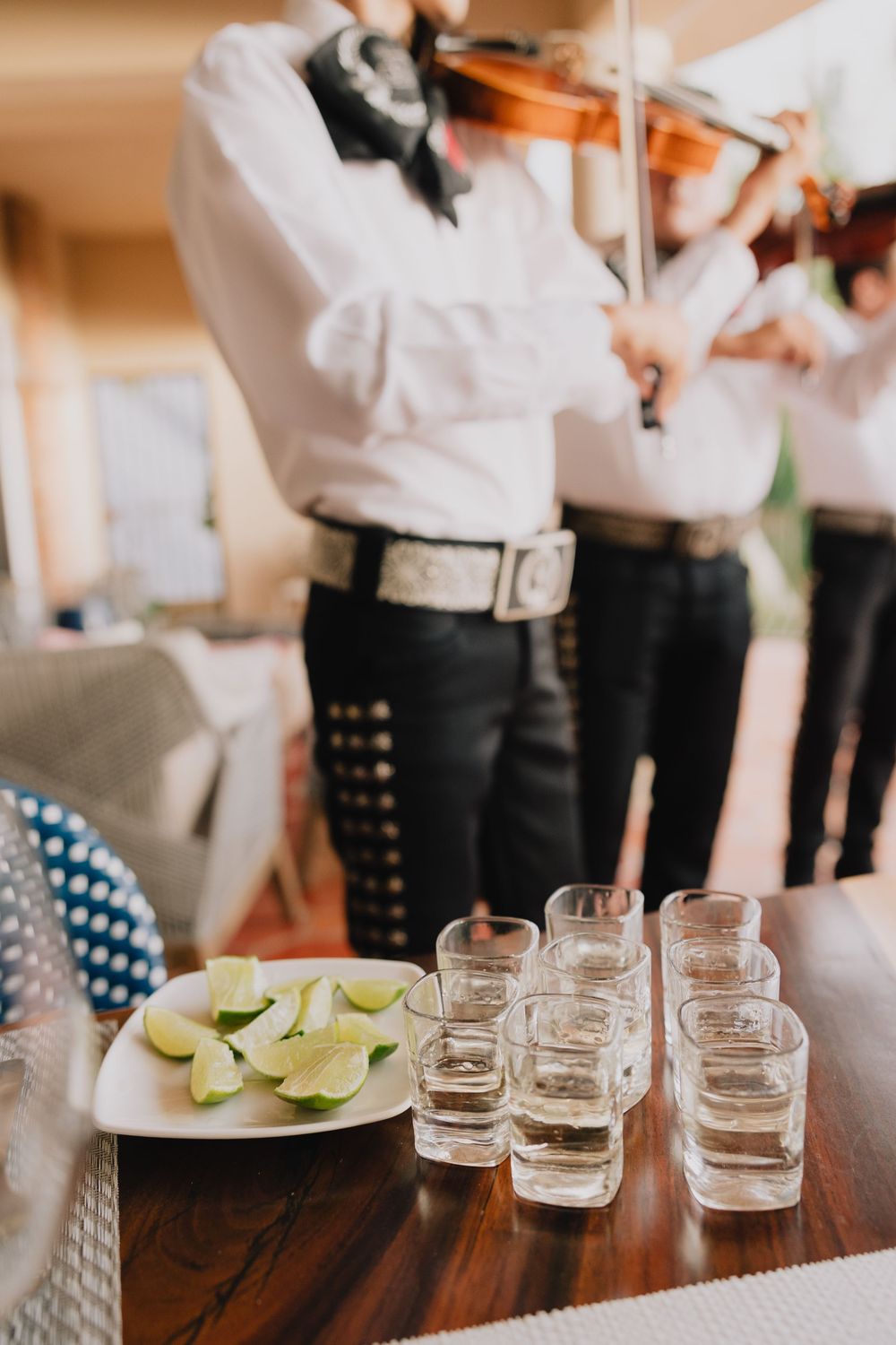 Tequila shots with lime wedges on a wooden table with mariachi musicians playing violin in the background.