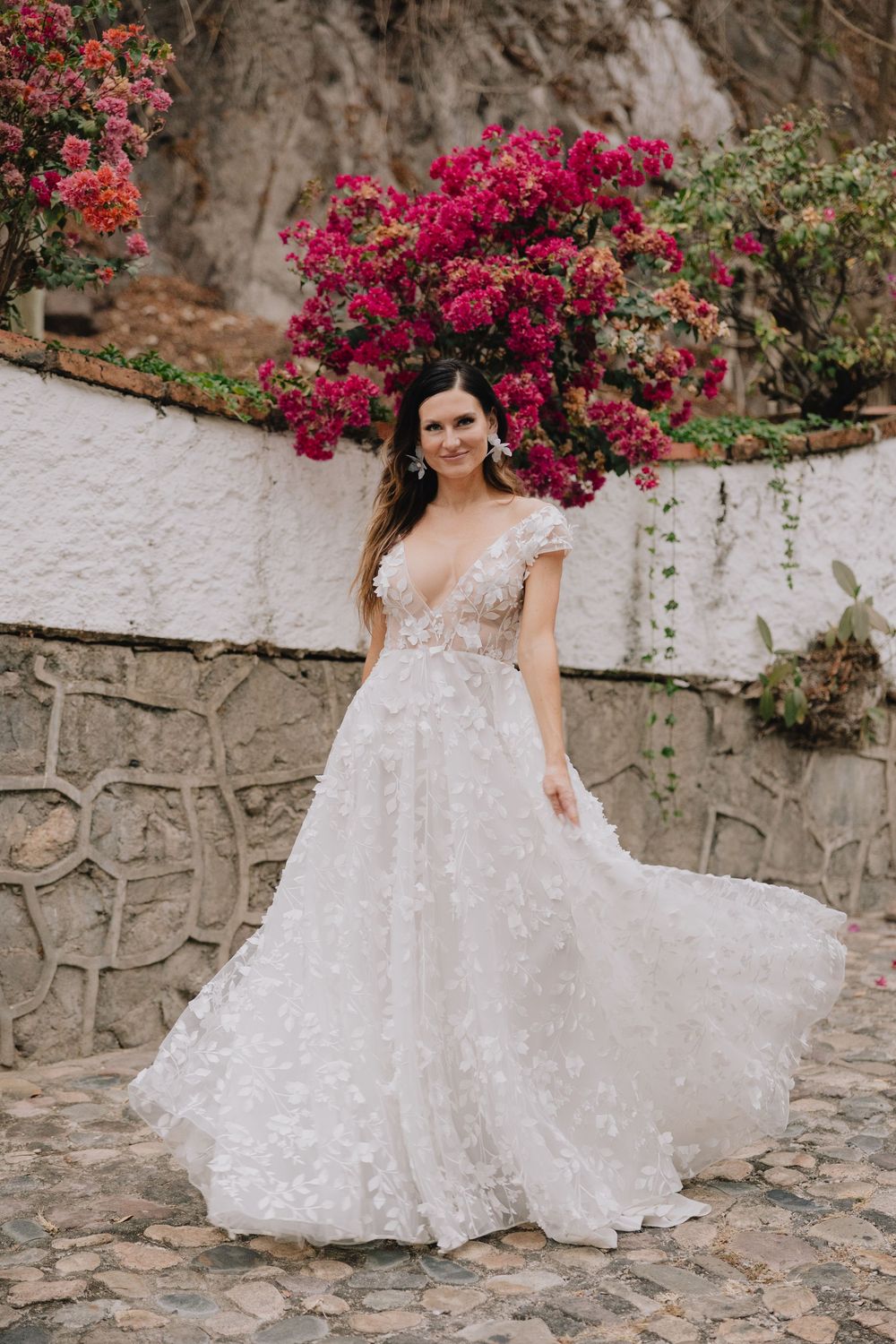 Bride in floral applique A-line wedding gown poses before vibrant bougainvillea blooms on stone wall.