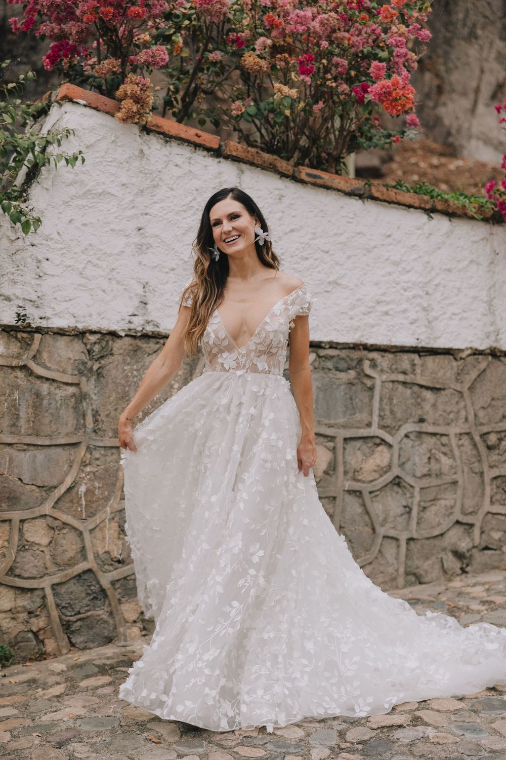Bride in floral lace A-line wedding gown smiling outdoors near stone wall with pink blooming flowers.