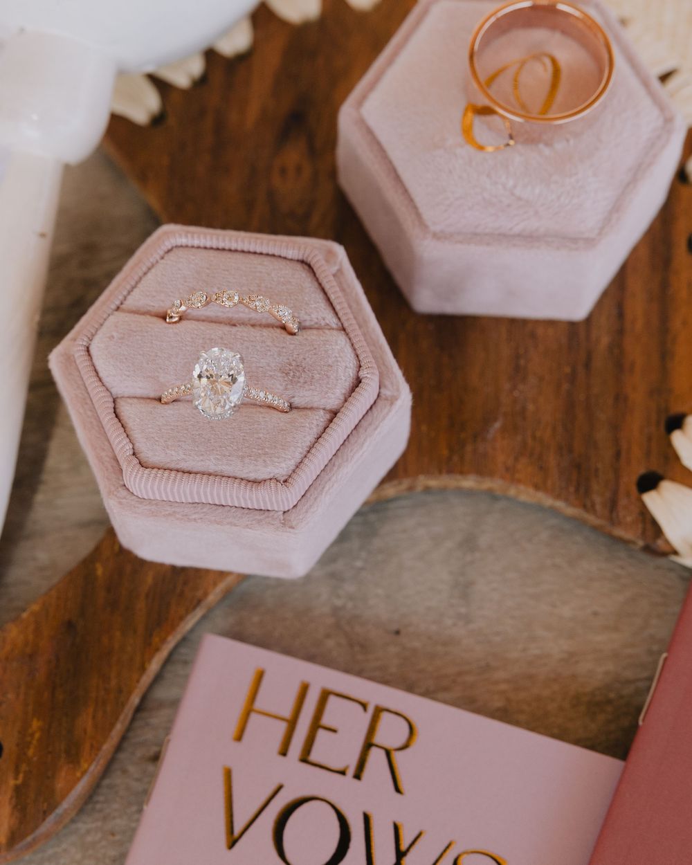 Oval diamond engagement ring and gold wedding band displayed in pink velvet hexagon ring boxes with vow booklet.