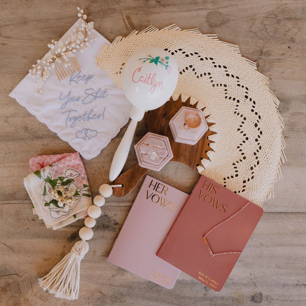 Flat lay of bridal accessories including vow books, a personalized mirror, ring box, and beaded tassel.