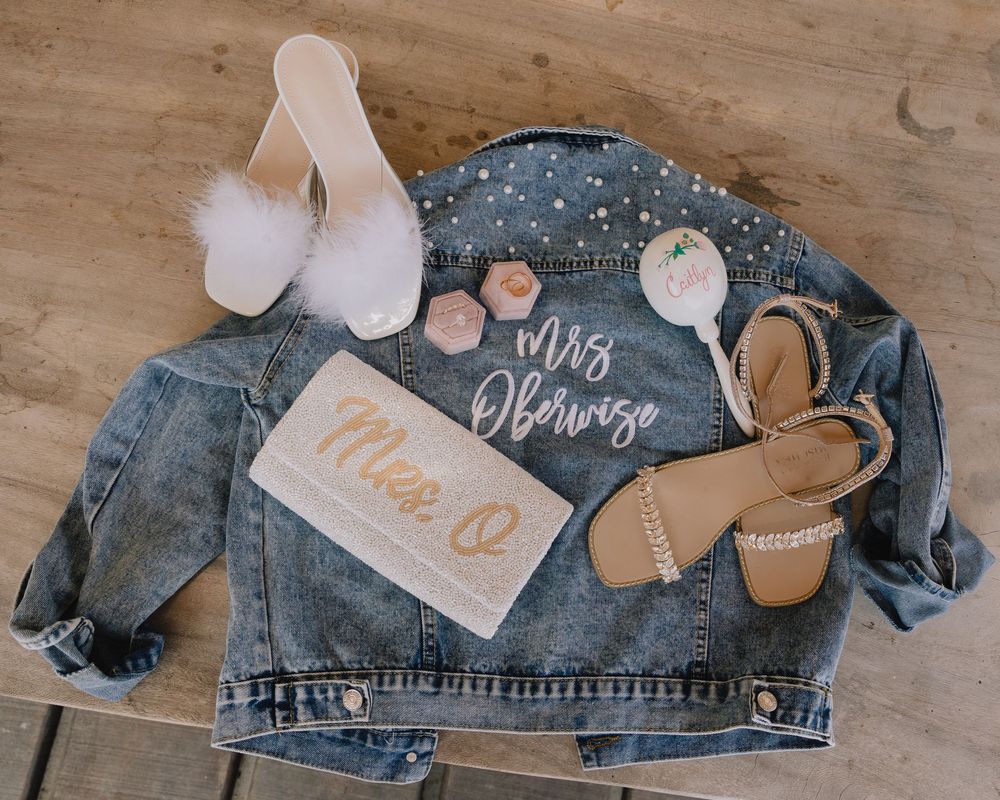 Bridal flat lay featuring pearl denim jacket, heels, sandals, and Mrs. Obariso accessories on wood floor.