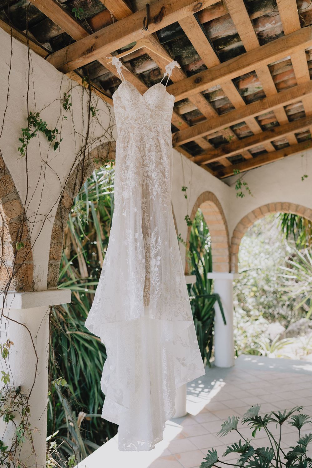 Elegant lace wedding dress hanging from a rustic wooden pergola surrounded by lush tropical greenery.
