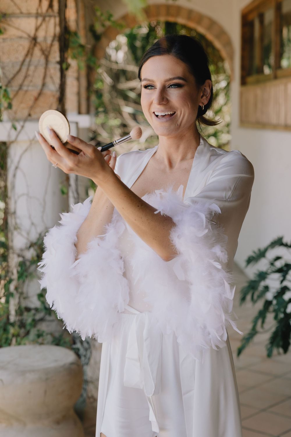 Happy bride in a white feather robe applying makeup with a brush while holding a compact mirror before her wedding.