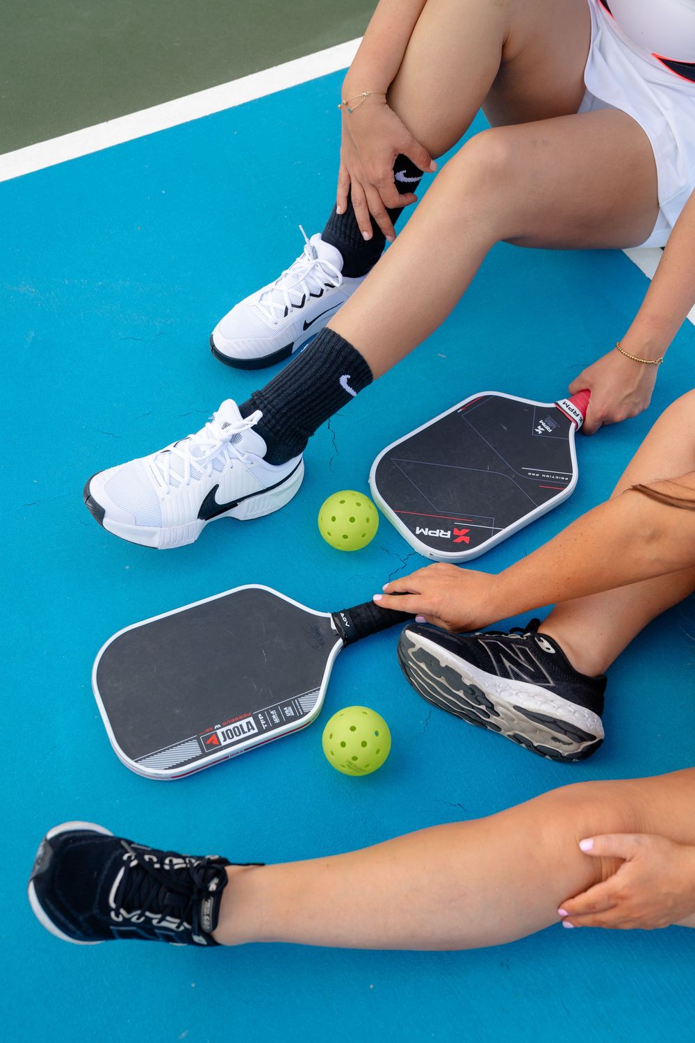 Close-up of pickleball paddles and balls on blue court with players' feet visible.