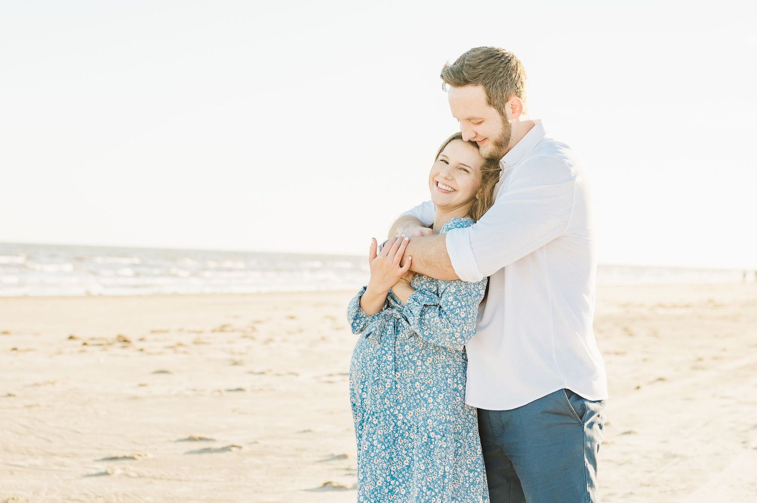 A couple embraces tenderly on a sandy beach during sunset, wearing blue and white outfits.
