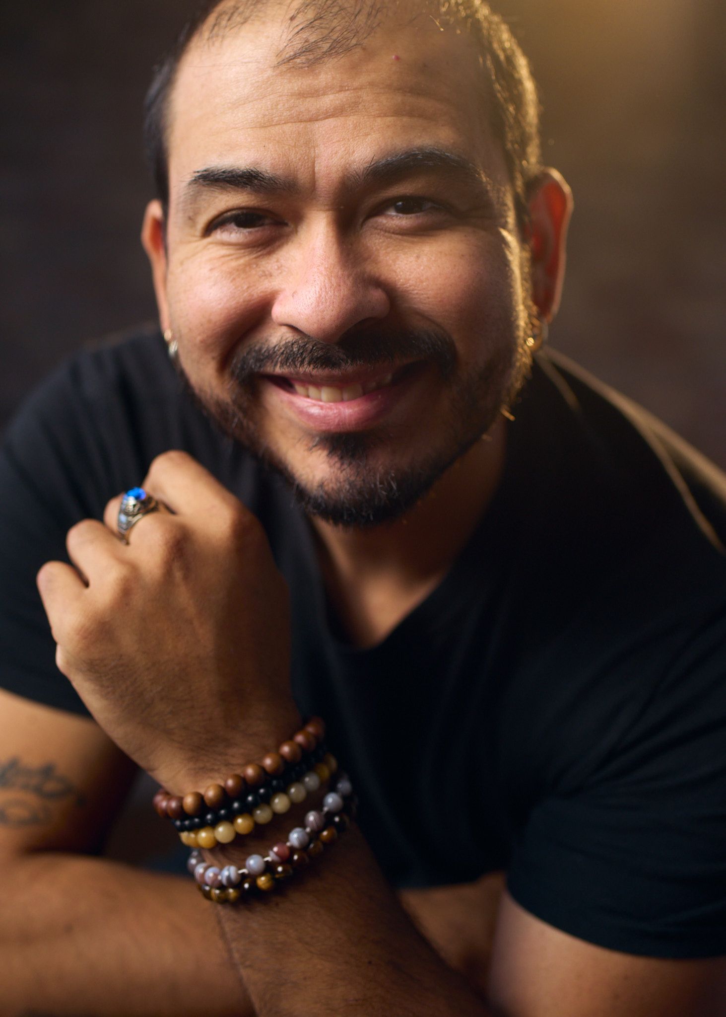 A smiling man with a beard wearing bracelets and a black shirt poses for a warm portrait photo.
