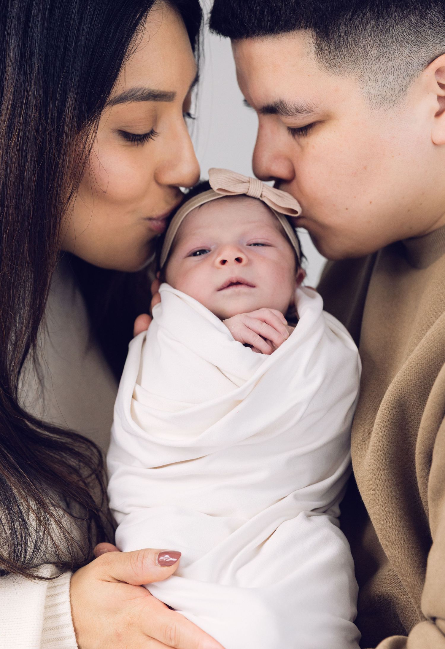Young couple kissing their swaddled newborn baby wearing a pink bow headband.