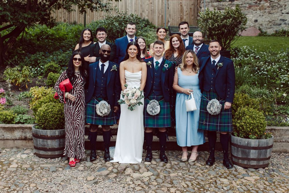 A group poses together for wedding photos in a garden setting with traditional Scottish kilts and formal attire.