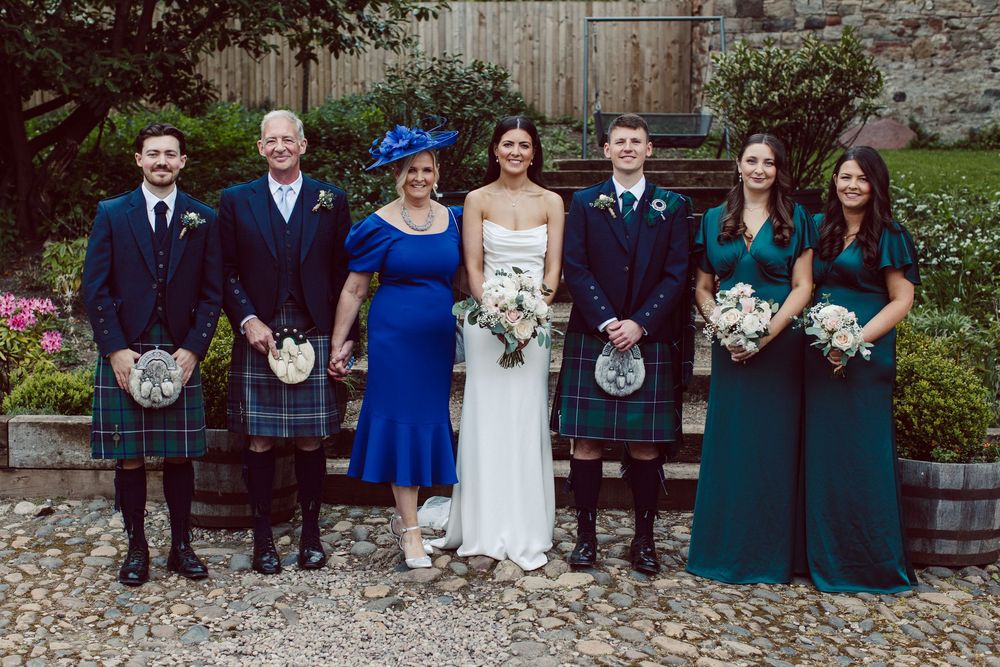 Wedding party members in royal blue and dark green attire sit together on a rustic wooden bench outdoors.
