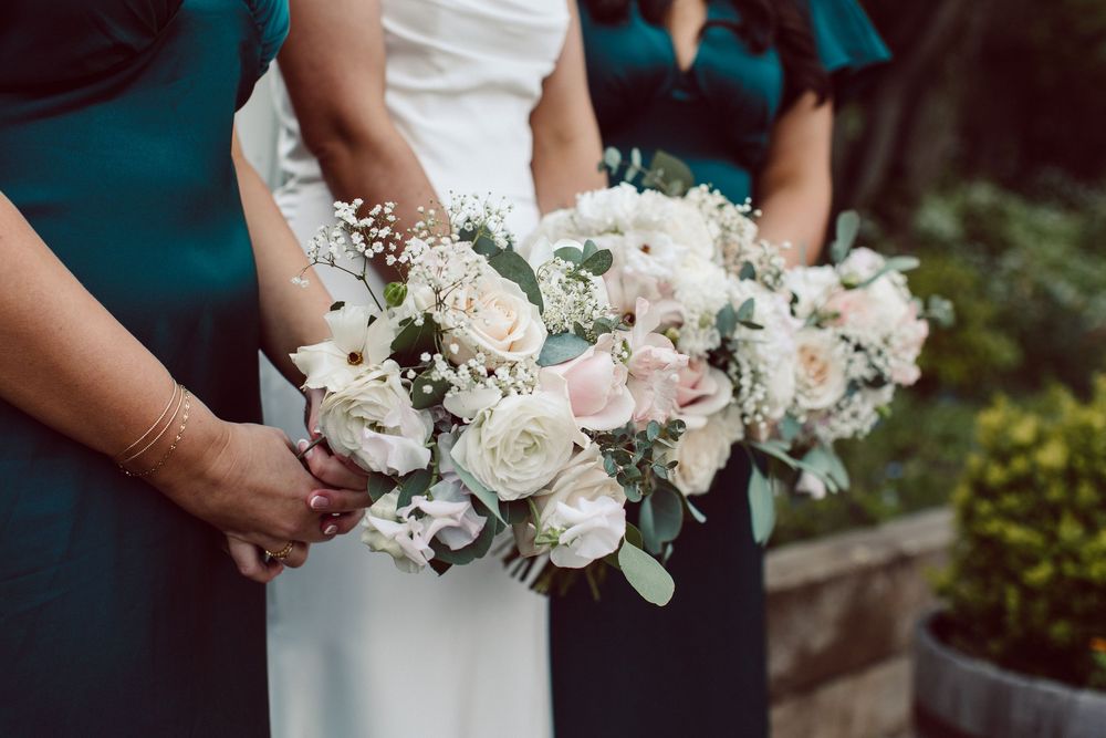 Wedding bouquets with white roses and greenery held by people wearing white and teal dresses.