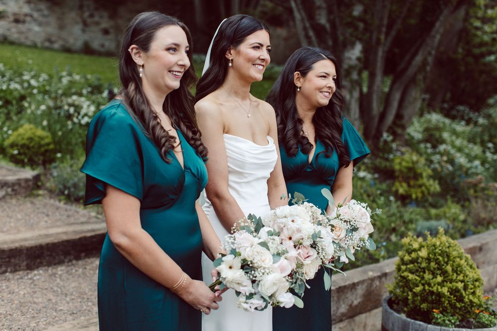 A bridal party in teal dresses and white bouquets shares joyful laughter during an outdoor wedding photoshoot.