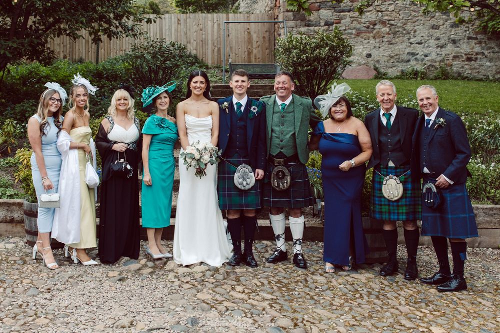 A wedding party poses for group photos in front of a stone wall in a garden setting.