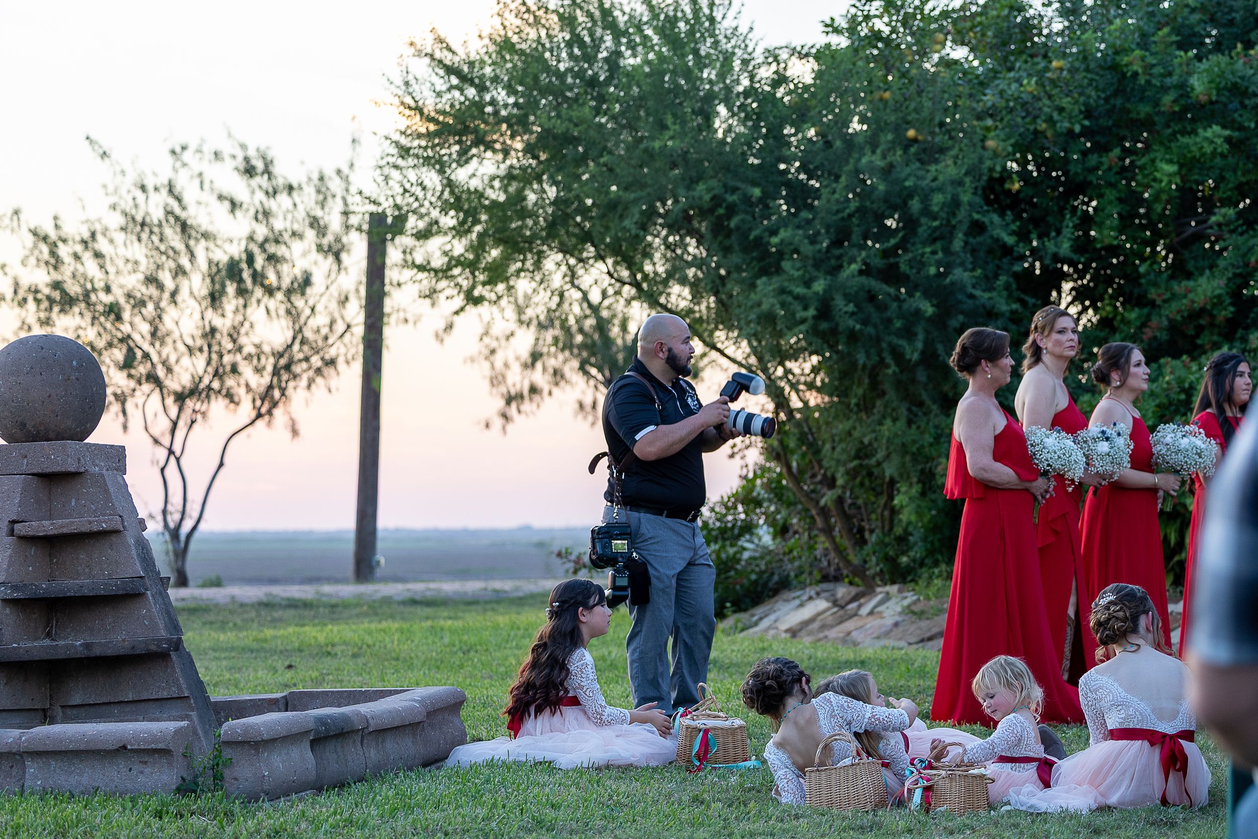 A photographer in black attire smiles while holding professional camera equipment outdoors at a rustic venue.