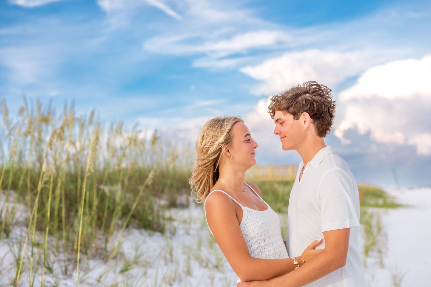 A couple shares romantic moments on a sandy beach with sea grass and blue skies in the background.