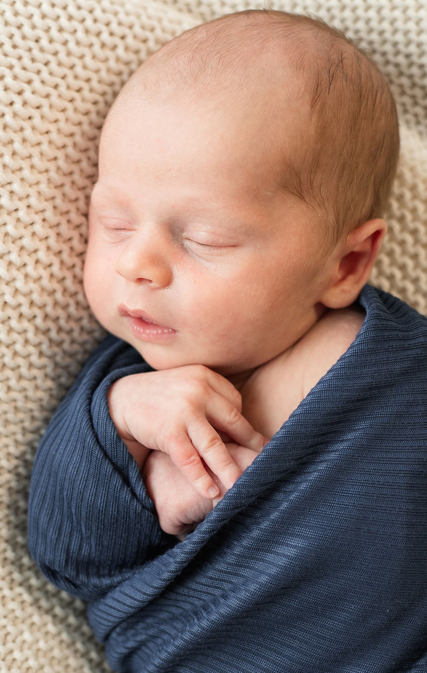 A sleeping newborn baby wearing a navy blue ribbed sweater rests peacefully against a cream-colored knit blanket.