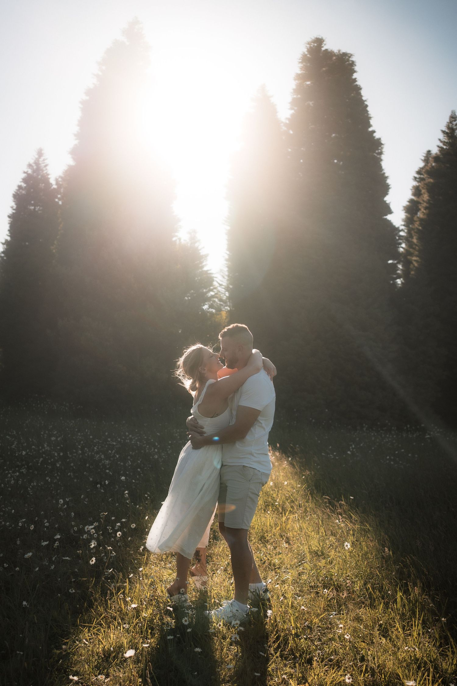 A couple embraces in a sunlit meadow during golden hour as sunbeams filter through evergreen trees in the background at RJ Hamer, Olinda