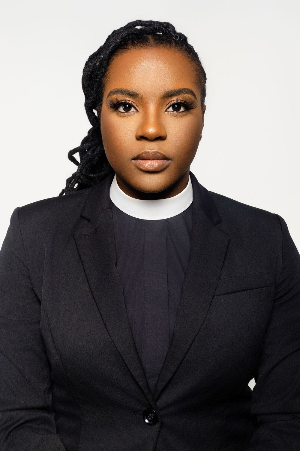 Professional headshot of someone in clerical collar and black blazer against white background.