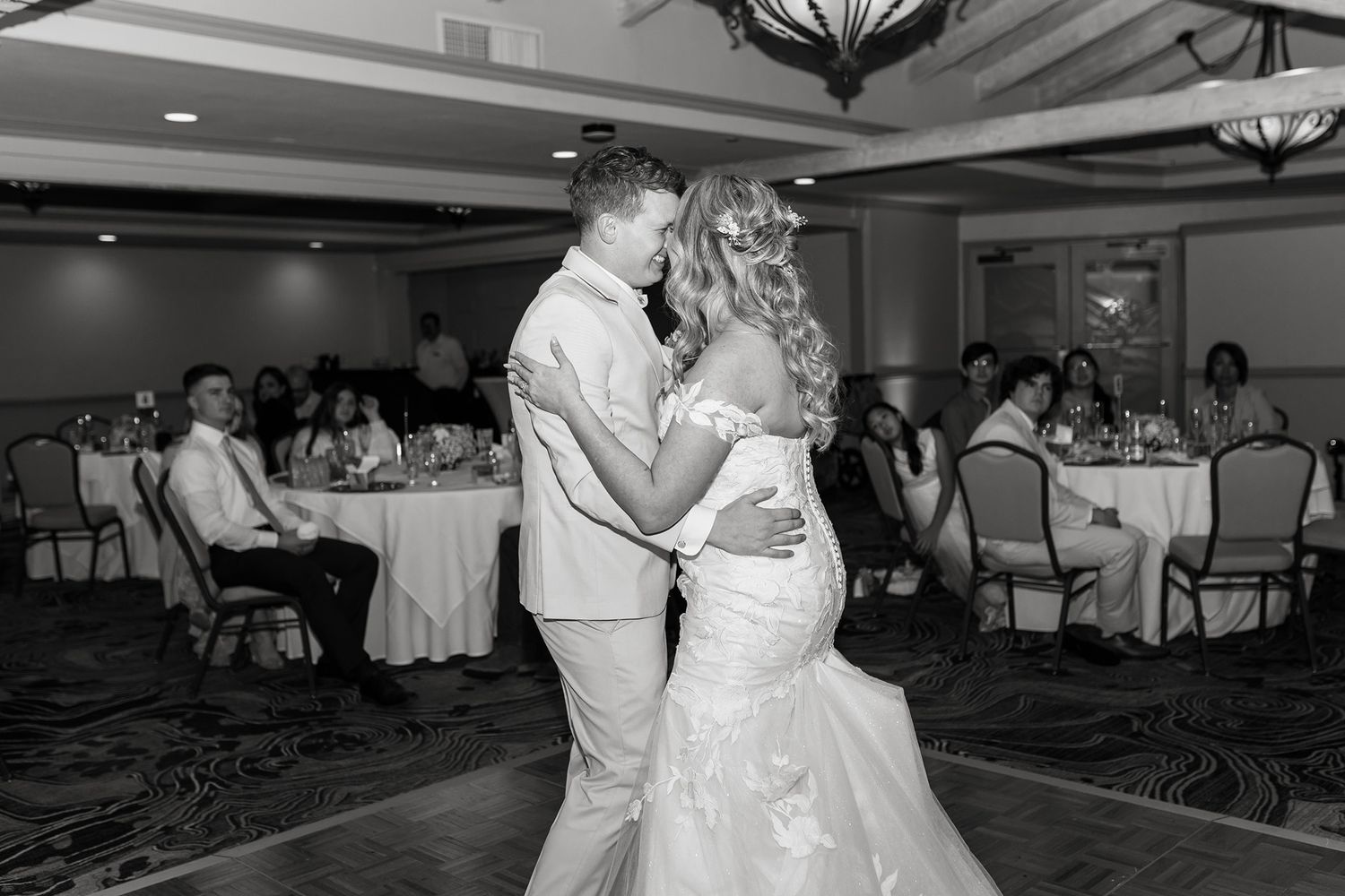 Couples dancing together at an elegant wedding reception in a ballroom with chandeliers and round dining tables.