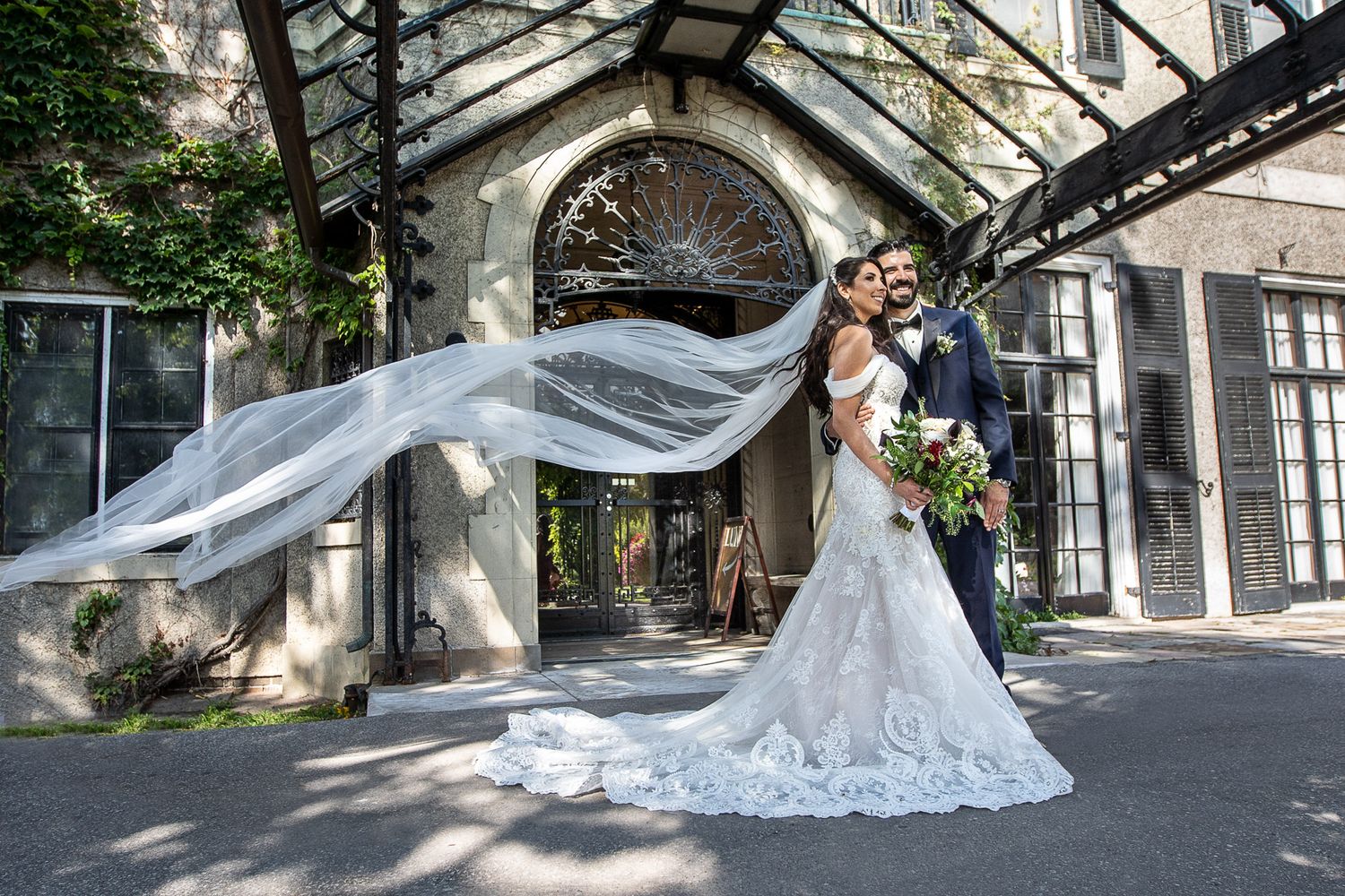 Bride and groom embrace outside elegant venue as cathedral veil flows dramatically in the wind.