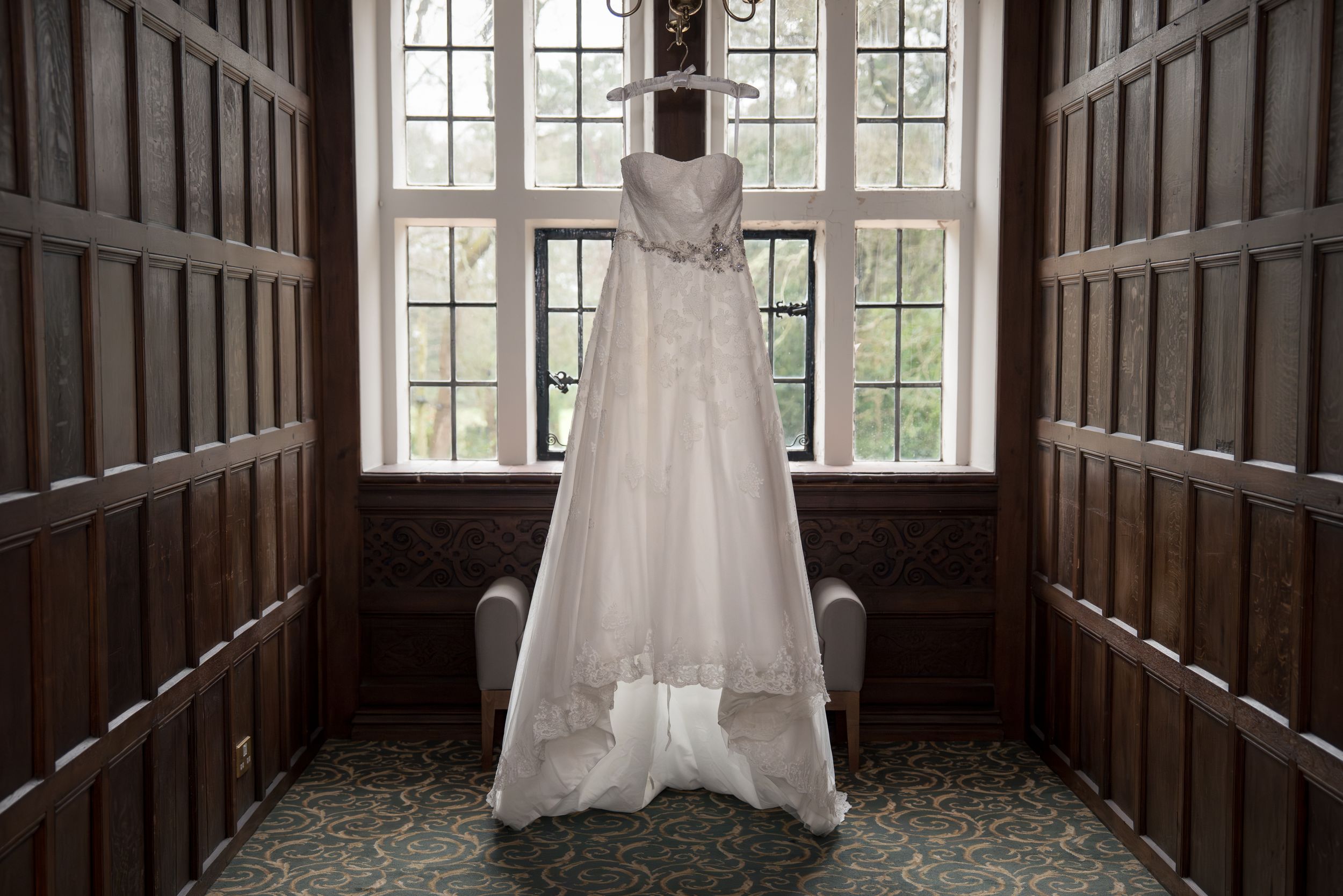 A white wedding dress hangs on display against a wooden paneled wall with a large window in natural light.