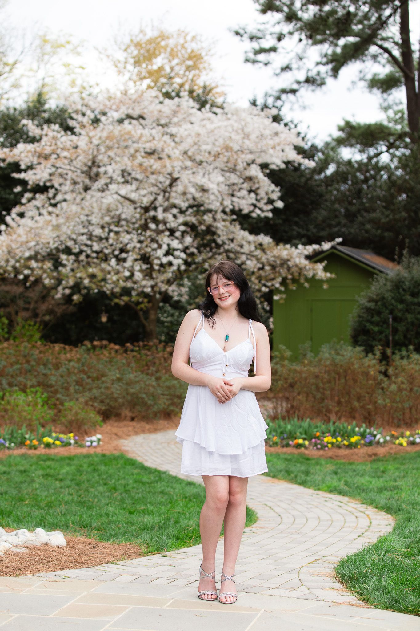 Senior Graduate in white dress standing on stone path with blooming white tree in spring garden at WRAL Azalea Garden