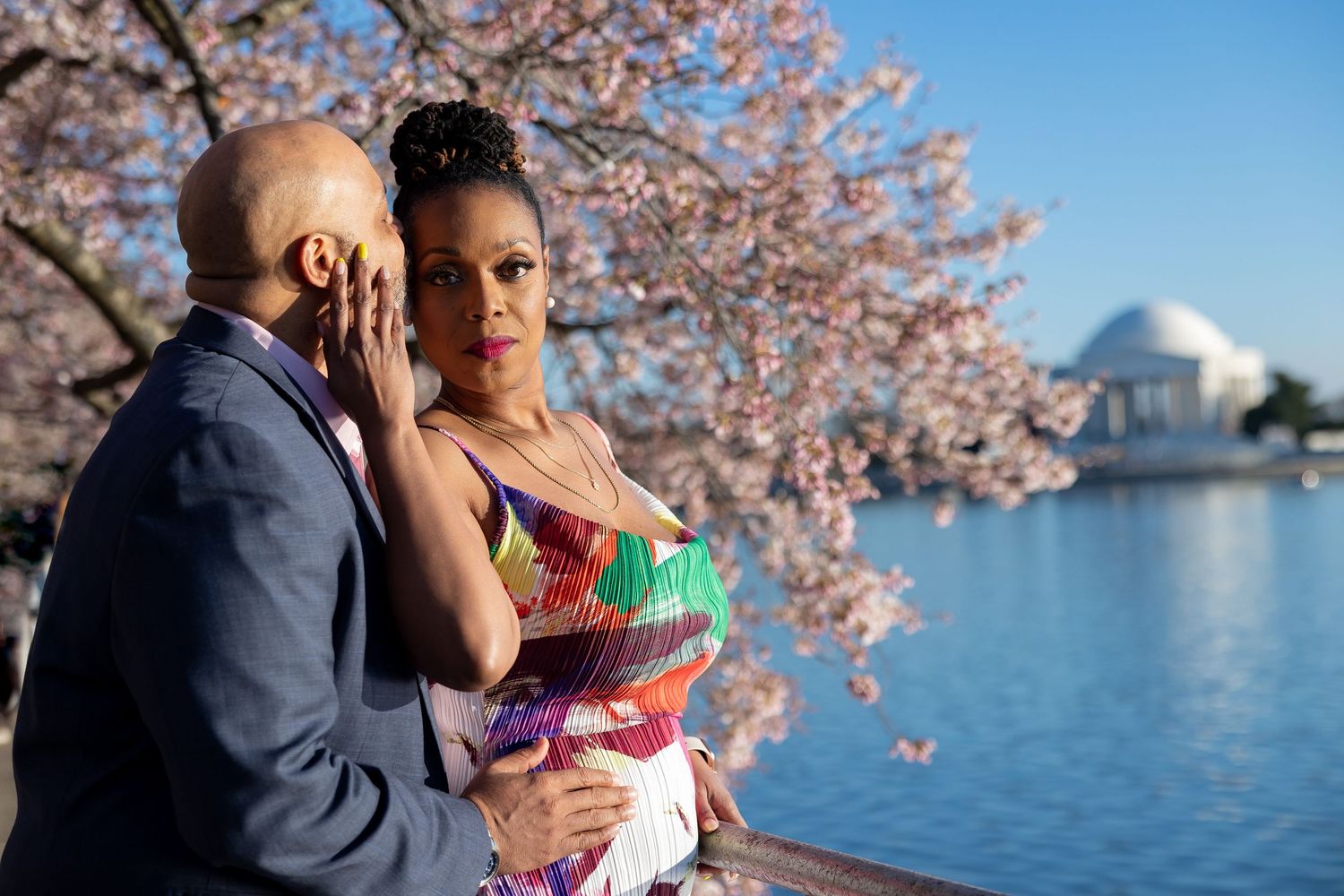 A couple embraces romantically near the Tidal Basin with cherry blossoms and the Jefferson Memorial in the background.