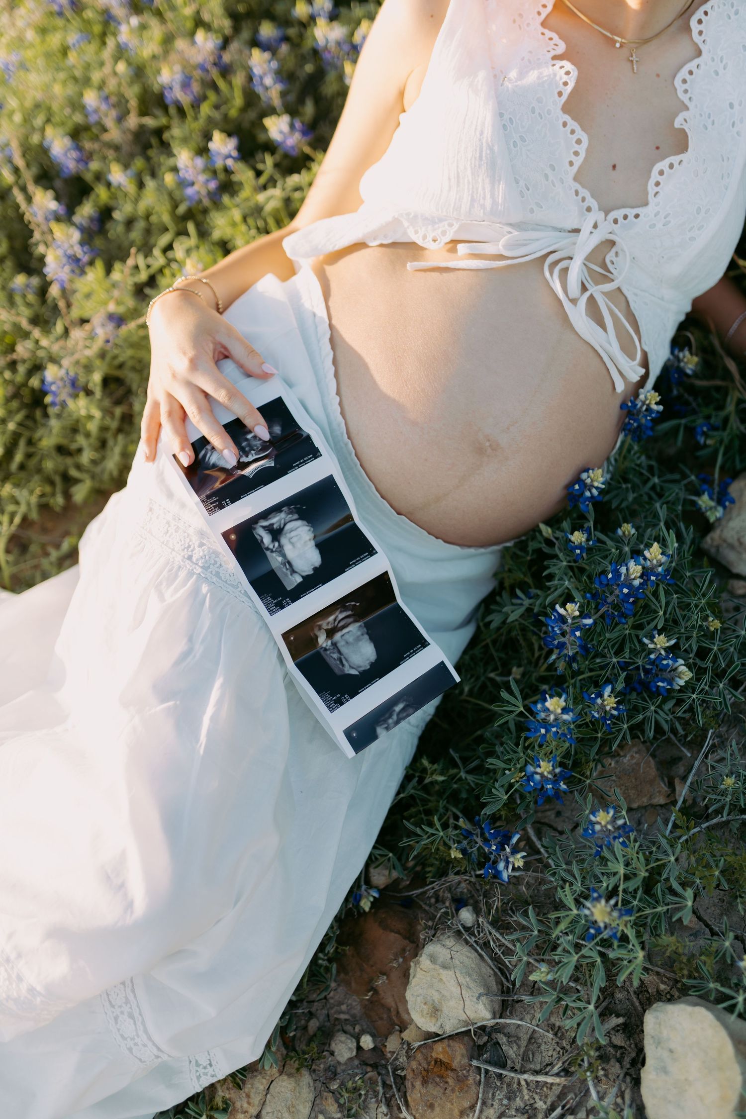 A person in a white dress holds ultrasound photos while sitting among blooming blue wildflowers in outdoor sunlight.