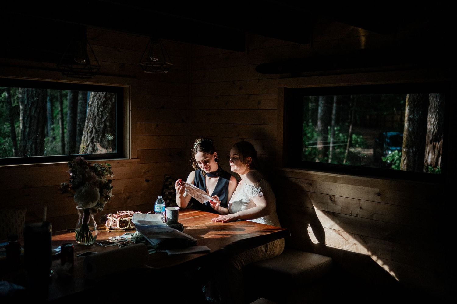 A dark and moody cabin interior scene with two brides gathered around a table illuminated by warm lighting.