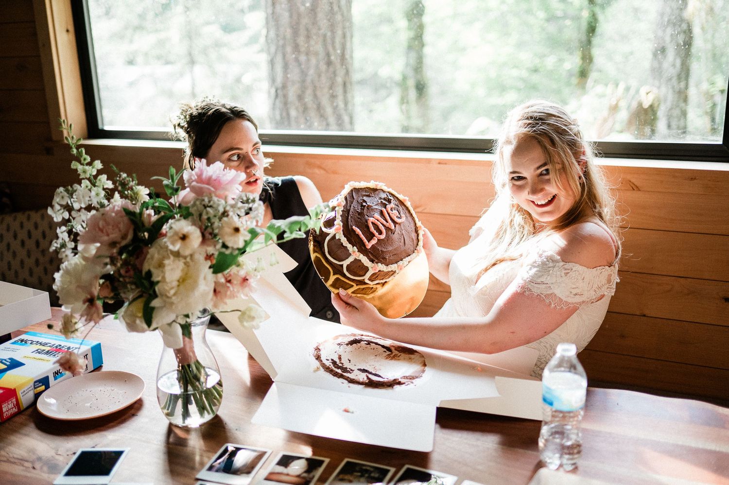 Two people share a joyful moment at a rustic wooden table with a floral centerpiece and cake plate.