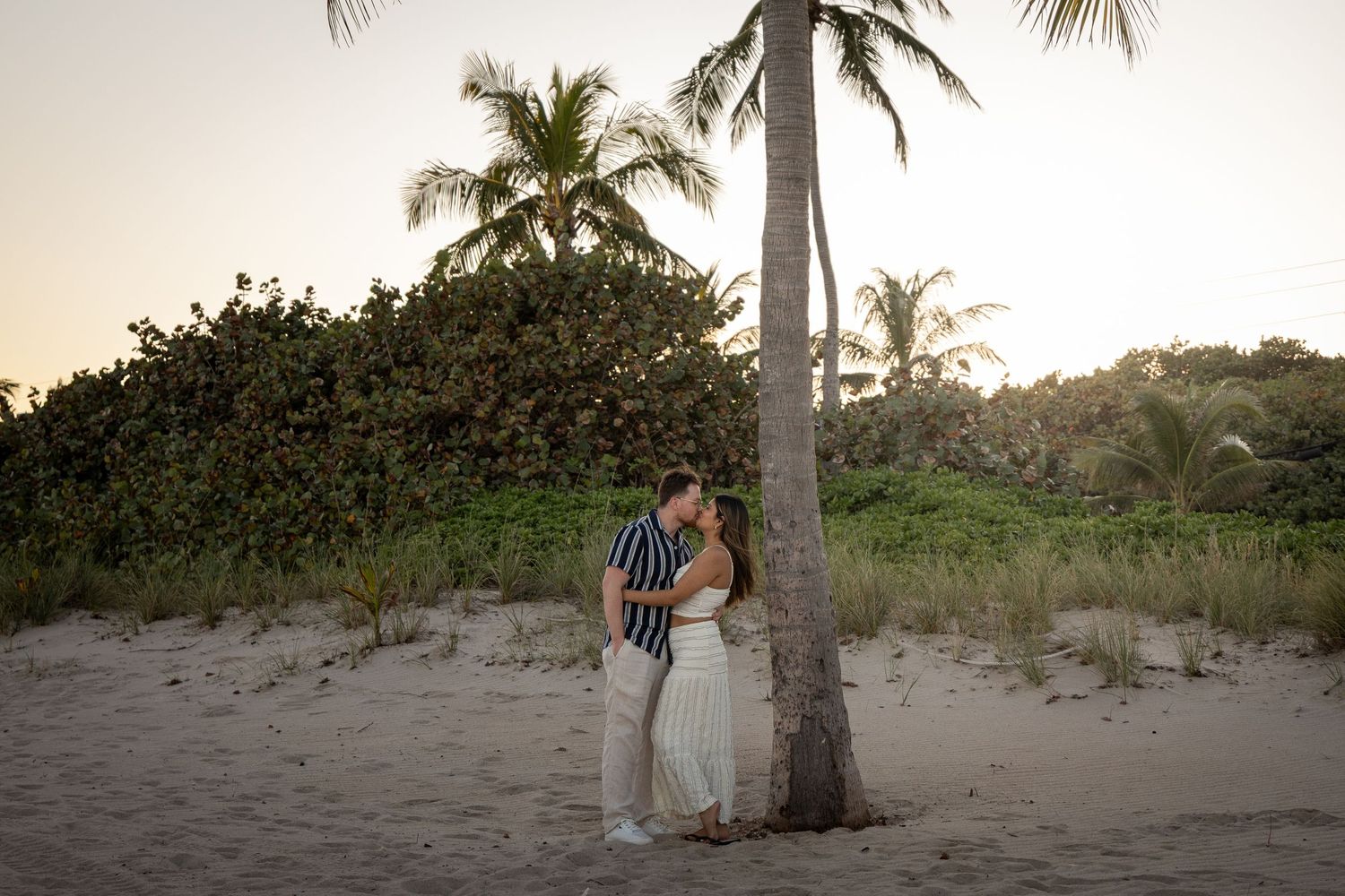A couple walks along a sandy beach path with palm trees and lush greenery at sunset.