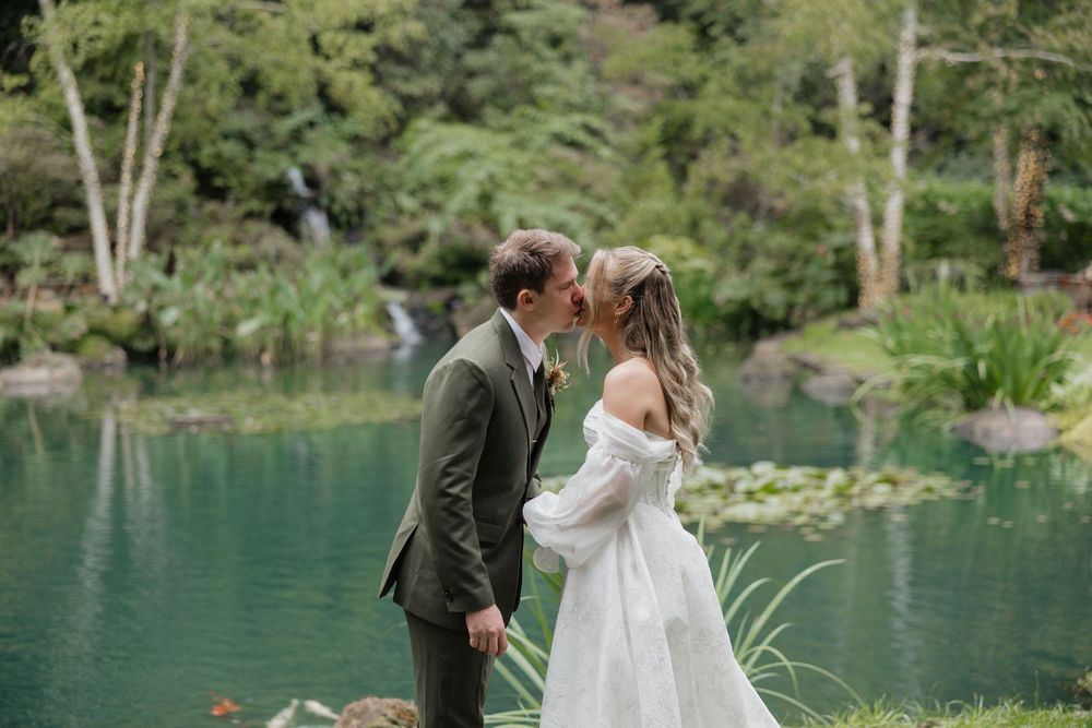 A couple shares an intimate first look moment beside a tranquil lake surrounded by lush greenery.