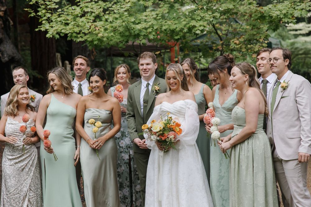 A wedding party wearing sage green dresses and light colored suits stands together outdoors for a group photo.