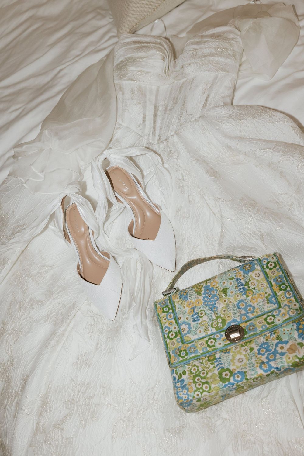 White wedding dress fabric with elegant white heels and a vintage floral print handbag arranged on light background.