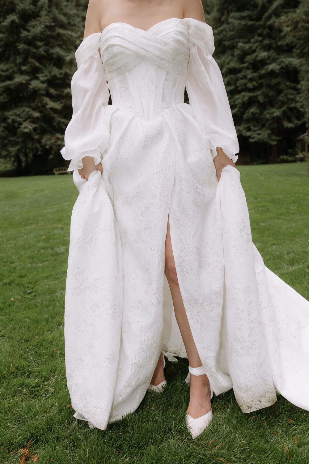 Detail shot of a white wedding dress with pockets and slit, showing white heels on grass.