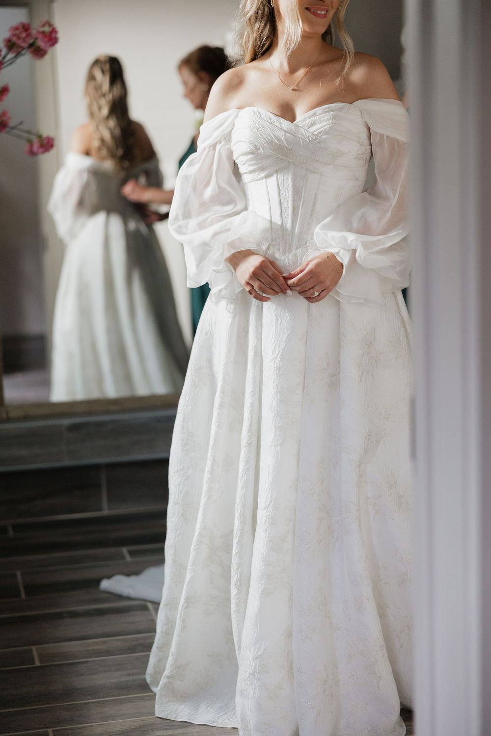 A bride in an elegant off-shoulder white wedding gown with billowy sleeves stands in front of a mirror reflection.