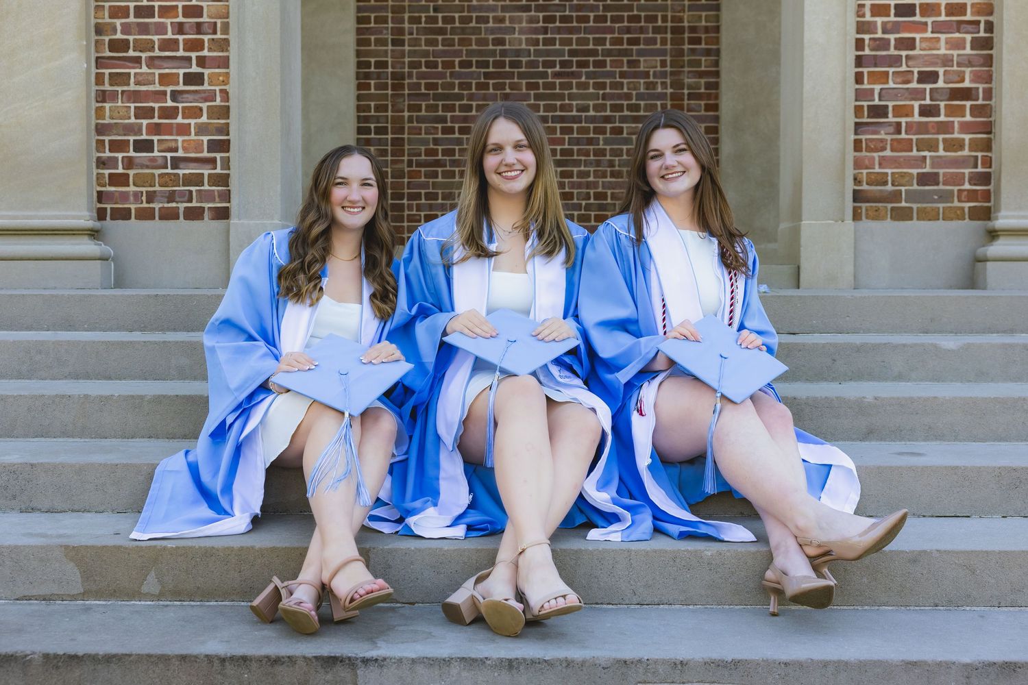 Three smiling women in blue graduation gowns sitting on stone steps outside a brick building.